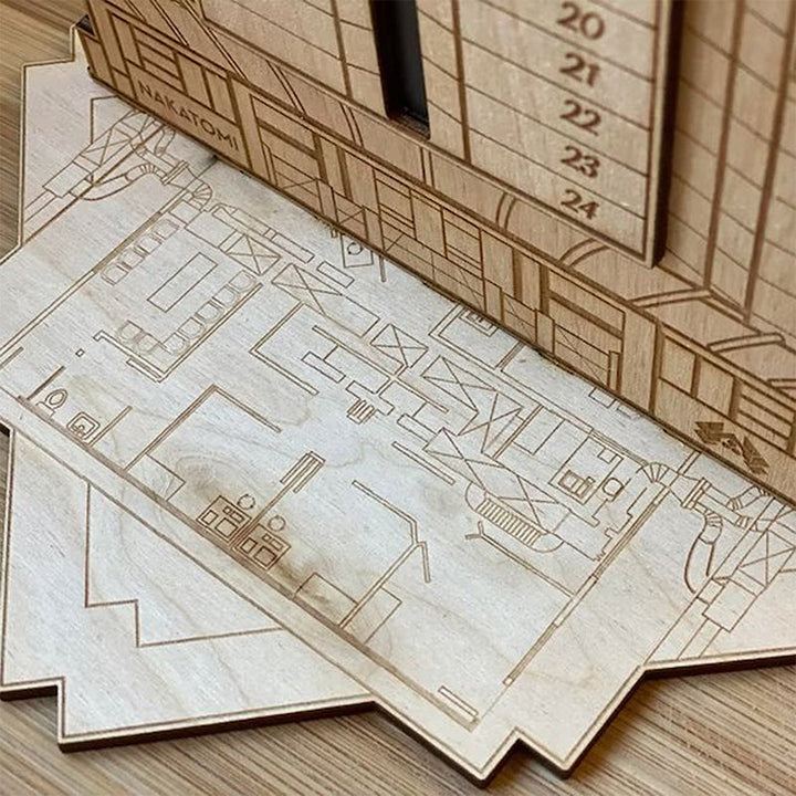 "Detaljerad trä Adventskalender med Die Hard-motiv, visar Hans Gruber faller från brunblå Nakatomi Plaza, roterbar design."