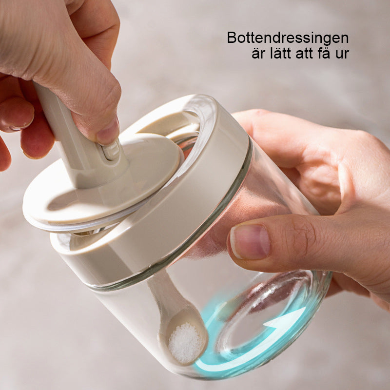 Kryddburk i glas med grönt och vitt skedlock, justerbart för enkel dosering, elegant design för köksanvändning.