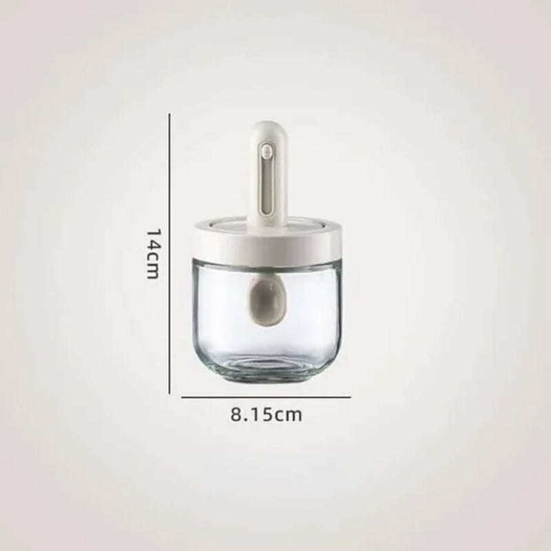 Kryddburk i glas med grönt och vitt skedlock, justerbart för enkel dosering, elegant design för köksanvändning.