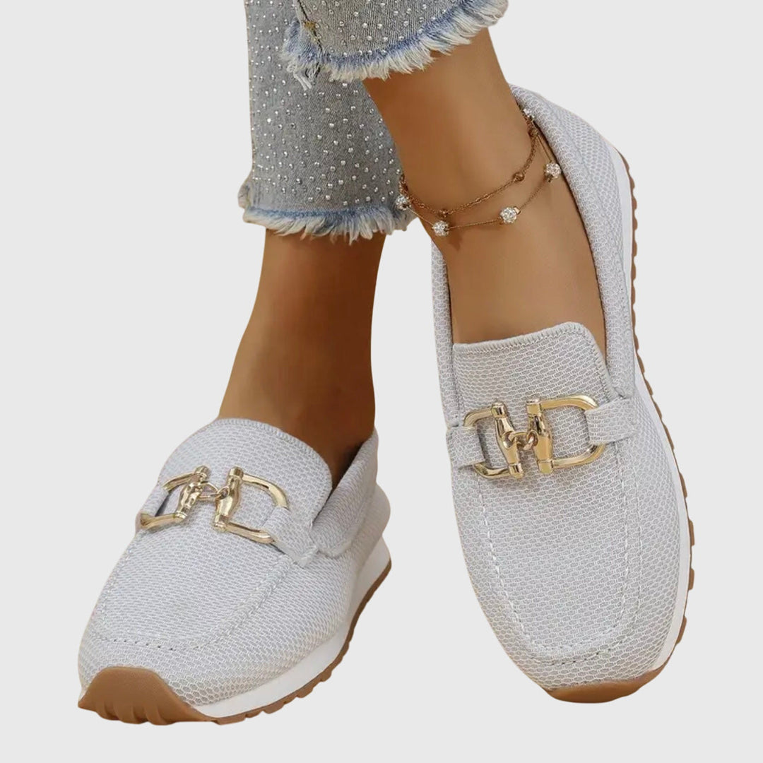 Två par stilrena slip-ons för dam, ett par vita och ett par beiga, på en trädäck.